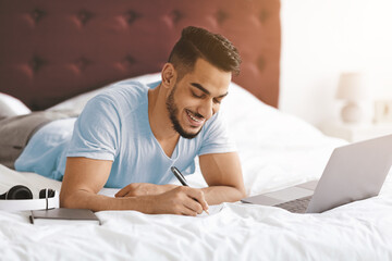 A young man is lying on a bed, using a laptop and writing notes. He looks relaxed and focused while enjoying his study session in a cozy room with warm lighting.