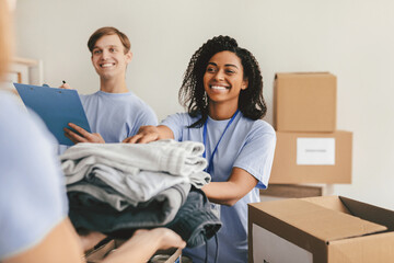 Two volunteers sort and pack clothing donations at a community center. One volunteer smiles while handing a neatly folded stack of clothes to another volunteer. It is a sunny afternoon.