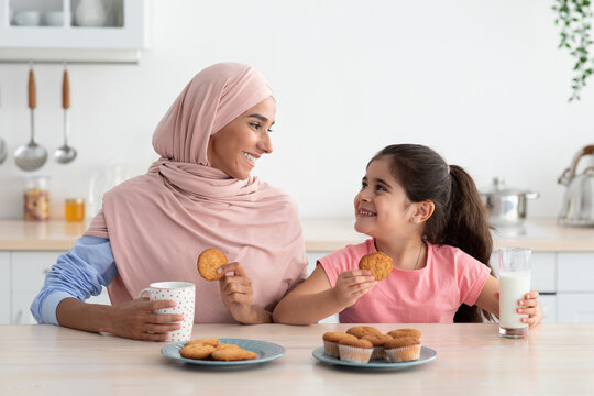 A mother wearing a headscarf shares a joyful moment with her daughter as they savor cookies and milk at a kitchen table. The afternoon sun fills the room with warmth.