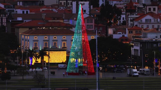 Aerial dusk view shows a cone LED Christmas tree in Funchal, Madeira. Color bands and musical notes cycle as tourists walk, buses load, and warm facades glow with lights.