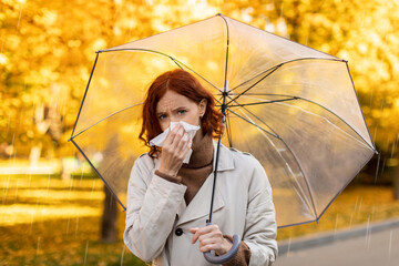Naklejka premium A woman with red hair is standing in a park, holding a tissue while she uses a clear umbrella for protection from the rain. Autumn leaves are falling around her, adding color to the scene.