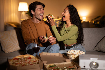 Happy spouses are sharing pizza and feeding each other as they relax on the couch. They are enjoying a cozy movie night together, creating warm memories and laughter at home.