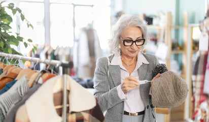 In sales area of store, mature female customer chooses color and style of winter hat. Creating capsule wardrobe. Casual practical clothes made of natural fabrics. Buyer enjoys shopping