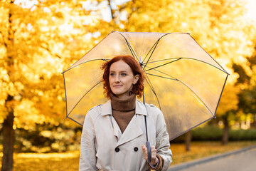 Naklejka premium A woman with red hair enjoys a walk in a park filled with yellow autumn leaves. She holds a clear umbrella while wearing a beige coat, smiling at the pleasant weather.