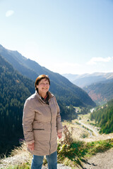 Active smiling senior woman, happy elderly traveler standing against scenic Fagaras mountains landscape. Transfagarasan Highway, Romania. Travel concept