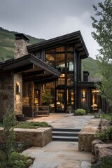 Modern house design located in a mountain area with large windows showcasing nature in the evening light and outdoor seating on a stone patio for relaxation