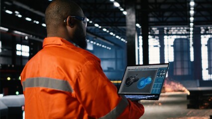 Technician in a manufacturing facility uses automation software on laptop to manage a turbine system surrounded by heavy equipment, steel machinery and fabrication tools.