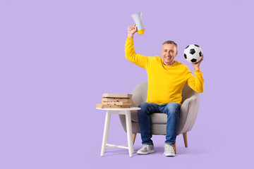 Male sports fan with soccer ball and megaphone in armchair on lilac background