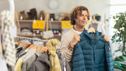 Stylish man standing in modern clothing shop and looking new down jacket made of high-quality...