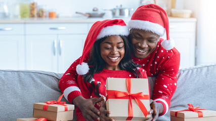 A couple in festive attire shares a joyful moment while opening a Christmas gift together on the couch. Surrounding them are neatly wrapped presents, adding to the holiday spirit.