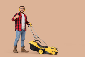 Young man in hearing protectors with lawn mower showing thumb-up on beige background