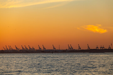 A vibrant sunset shows an industrial container seaport in New York with silhouettes of cranes.
