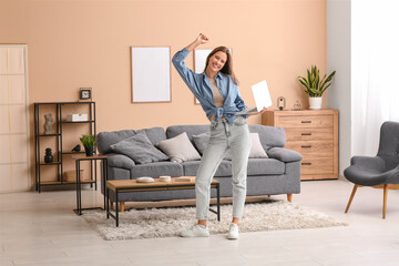 Beautiful young woman dancing with modern laptop in living room