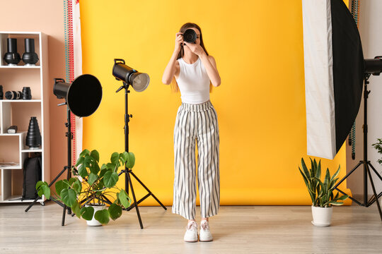 Female photographer with camera and houseplants near yellow backdrop in photo studio - Powered by Adobe