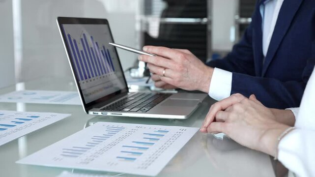Business professionals collaborating in a modern office, reviewing financial reports, charts and a laptop on a glass table while discussing data analytics, strategy and planning