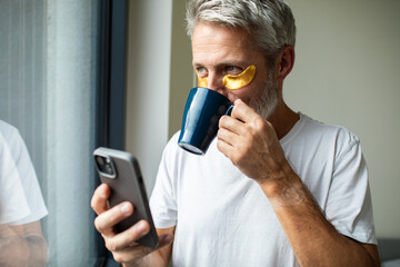 Mature man with eye patches drinking coffee, focused on smartphone at home