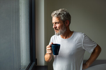 Mature man sipping coffee, looking thoughtful by window at home