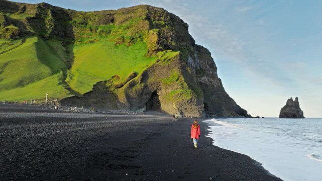 Aerial drone view of a woman walking on a black sand beach, Reynisfjara, Iceland. 4K cinematic footage highlights volcanic cliffs and Atlantic coastline.