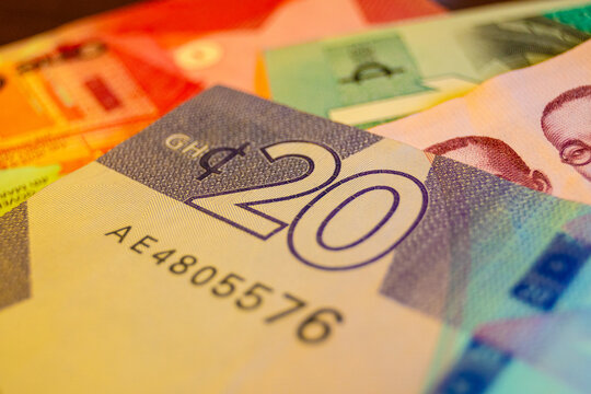 A close-up, shallow-focus shot of a 20 Ghana Cedi banknote showing the "20" denomination and security features. The purple note is layered over other blurred Ghanaian currency in a warm light.