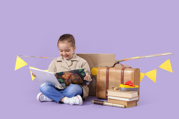 Fototapeta premium Cute little girl with cardboard plane and suitcase reading adventure book on lilac background