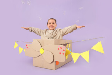 Fototapeta premium Cute little girl in cardboard plane with soap bubbles on lilac background