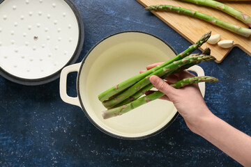 Female hand with fresh green asparagus and cooking pot on blue background