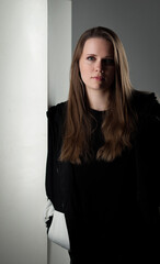 Studio shot of a beautiful young woman with long brown hair wearing a black jacket leaning against a white wall. This image is suitable for fashion beauty and lifestyle concepts.