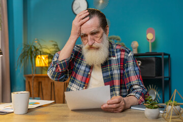 Shocked senior man freelancer analyzing paperwork graphs, charts, accounting payments and calculating monthly expenses bank bills at home office desk. Upset sad grandfather working remote job at table
