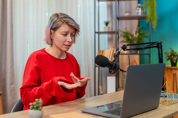 Woman recording video interview at laptop sitting at home table, speaking seriously in studio microphone. Caucasian girl confidently looking at webcam, professional expression demonstrating expertise.