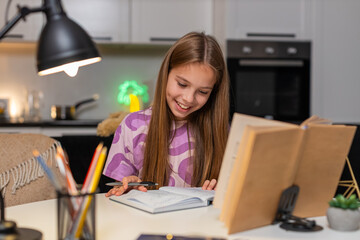 Teenager girl at night home works on homework with book writes notes in notebook stays serious. Caucasian child kid sitting at table at evening solves math task checks example holds pen writes result.