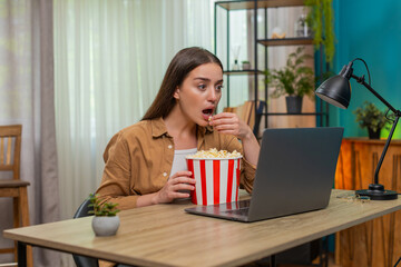 Young woman sitting at home table watches dramatic film on laptop eating popcorn and feeling tense. Caucasian girl grips seat nervously reacting to intense plot twists while chewing popcorn quickly