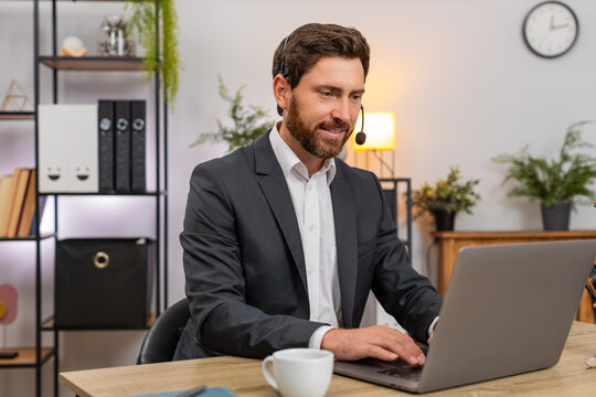 Fototapeta Middle-aged businessman at home office works as call center operator with headset listens and types solution on laptop for client. Freelancer guy at table speaks kindly confirms steps and saves notes.