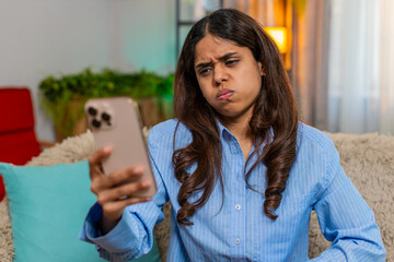 Indian woman reads bad news on smartphone and sighs sadly while sitting on home sofa, lowering eyes in disappointment. Arabian girl expresses quiet sorrow, negative emotion, concern and fatigue.