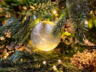 The image shows a close-up of a festive Christmas tree branch with a delicate, frosted glass bauble hanging among the green needles, illuminated by soft golden lights, typical symbol  of Christmass