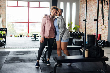 African American female friends standing in gym wearing activewear and headphones by dumbbell rack