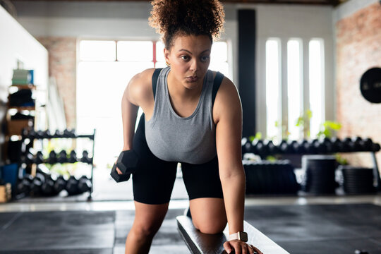 African American woman performing single-arm dumbbell row on bench in gym with dumbbell racks