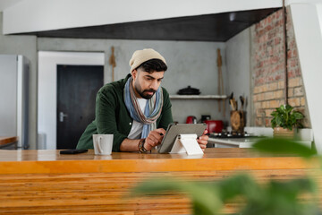 Indian man wearing beanie leaning at kitchen counter, using tablet stand beside mug, smartphone