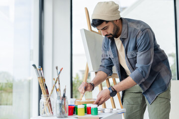 Indian male wearing beanie, denim shirt mixing paint on palette at art studio with wooden easel