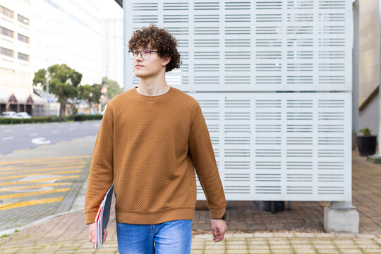 Male student walking on urban sidewalk beside metal wall holding notebooks and wearing eyeglasses