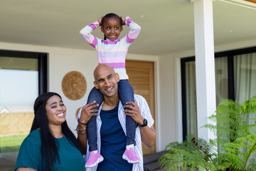 Diverse family standing on front porch carrying daughter on shoulders by woven wall hanging