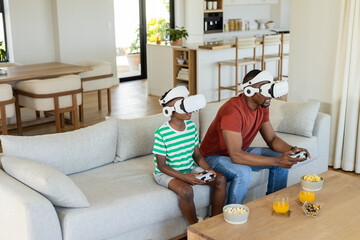 African American father and son using VR headsets and controllers on light-gray sofa at home