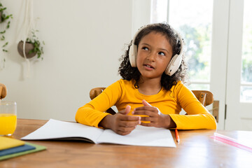 Child girl sitting at table in home dining area wearing yellow shirt and headphones using notebook
