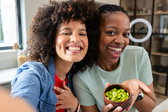 Female friends taking selfie in kitchen by ring light while holding wooden bowl of edamame beans