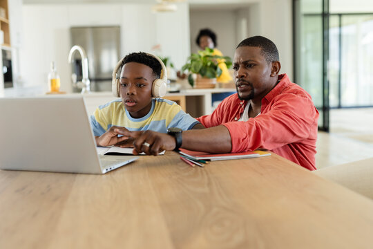 African American father and son working on laptop and notebooks at kitchen desk with headphones