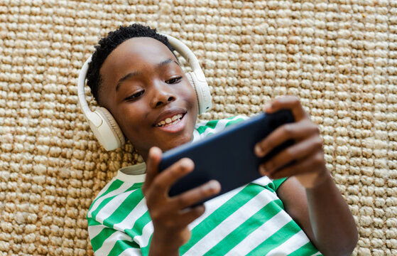African American boy lying on beige rug at home wearing white headphones and grinning at smartphone