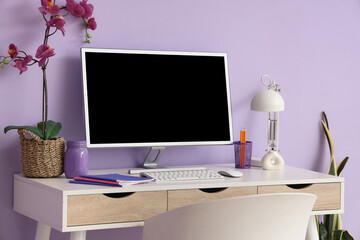 Workplace with blank computer monitor and orchid flower near lilac wall in office