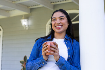 Smiling Asian woman leaning against white column holding terracotta ceramic mug on front porch