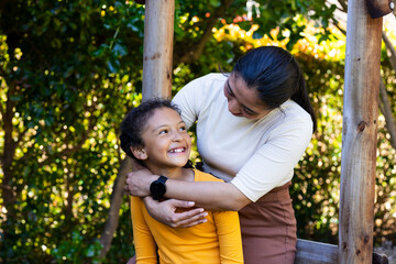 Hispanic mother hugging daughter standing behind bench under frame in backyard wearing smartwatch