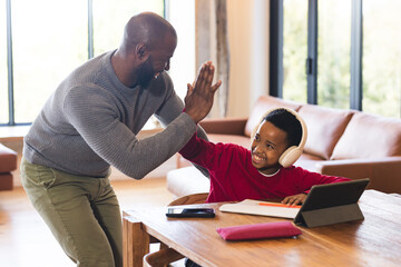 African American father leaning at table high-fiving son using tablet with headphones at home