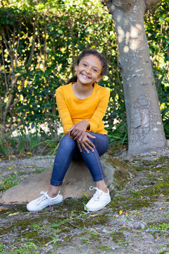 Female child sitting on stone near tree-trunk in park wearing yellow top, sneakers smiling in sun